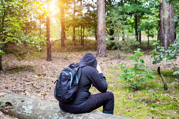 A man with a backpack in a hood sits on a fallen tree in the middle of a clearing in the forest....