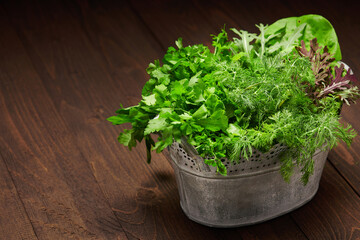 a bunch of green dill, parsley, salad and other greens in an iron bucket, dark wooden background, concept of fresh vegetables and healthy food