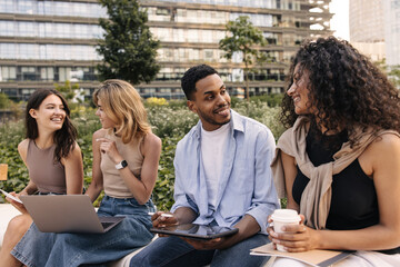Diverse young students are developing spreadsheet based on lectures laptop and tablet sitting outdoors. Guy and girls communicate on educational topics. Technology concept