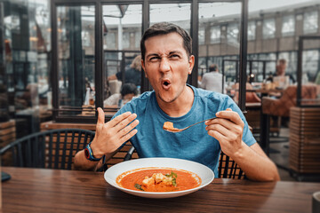 A man tries a spicy and hot red soup in a restaurant and reacts funny emotionally. Seasonings in the national cuisine and an unhealthy diet with overabundance of pepper - obrazy, fototapety, plakaty