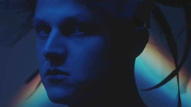 Portrait shot of man with nose piercing wearing veil and feathers on his head posing for camera in dark studio with blue neon light