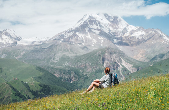 Backpacker woman sitting on a green grass hill and enjoying snowy slopes of Kazbek 5054m mountain with a backpack while she walking by green grass hill. East Caucasus mountains, Georgia..