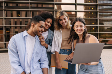 Young interracial students are doing group project on laptop sitting on campus. Guy and girls wear casual clothes in spring. Concept of learning.