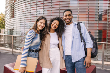 Friendly young interracial guy and girls embracing posing looking at camera spending leisure time on street. Brunette friends wear casual clothes for walk. Concept of enjoying moment