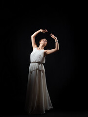 Fototapeta premium An ancient heroine, a young woman in the image of an ancient Greek goddess or muse. A noble heroine in a white tunic and a laurel wreath, dancing on a black background