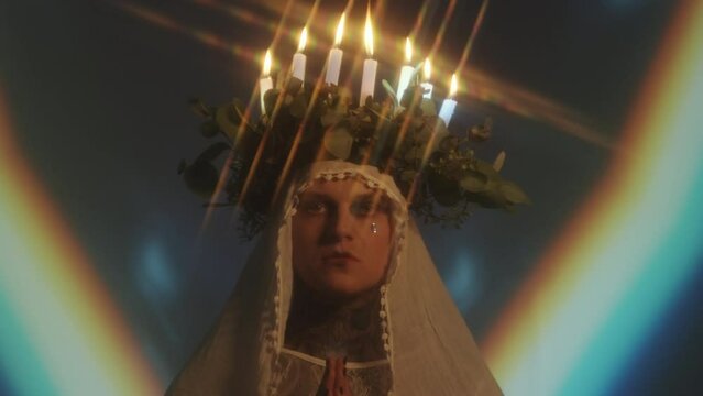 Portrait of man with candle lit wreath on his head, veil and face gem posing for camera with hands folded in dark studio with rainbow light projection in background