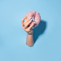Sweet dessert, a donut in pink glaze in your hand. beautiful woman's hand with a manicure. Hand gesture in a cardboard hole, blue background