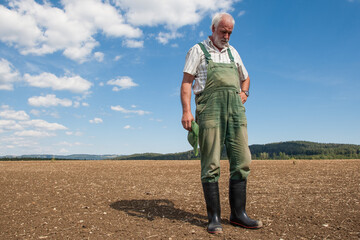 An experienced farmer in green dungarees and black rubber boots is standing in the freshly sown...