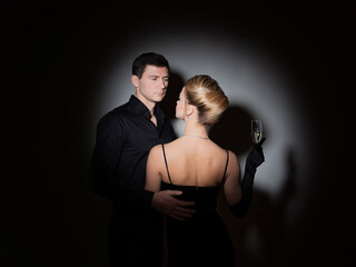 An attractive couple in black. Stylish and elegant young man and woman in the spotlight. Beautiful blonde in a long dress with a glass in her hands