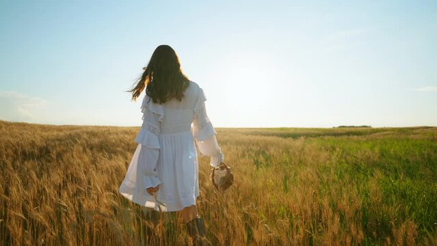 carefree brunette woman in white dress in country style is walking on field with golden rye or wheat