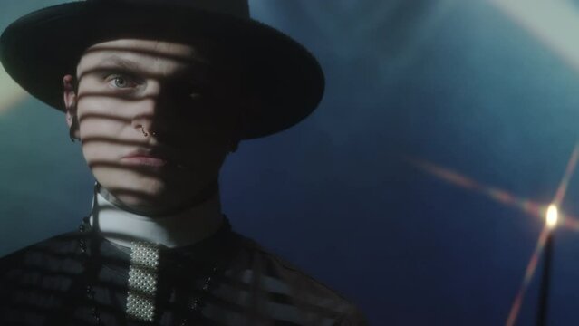 Chest up shot of young priest in cassock and hat standing with candle in studio with light projection