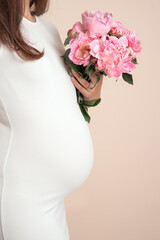 A body portrait of a young white pregnant female in white dress holding pink peonies on beige colored background