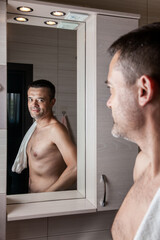 Fototapeta na wymiar A man looks in the bathroom mirror in the morning with a towel on his shoulder