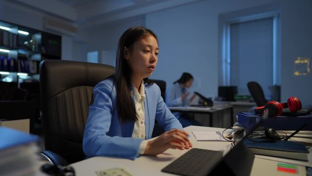 Stressed overburdened Asian woman having panic attack as angry Caucasian colleague arguing at background. Portrait of anxious worried employee breathing in paper envelope sitting in office