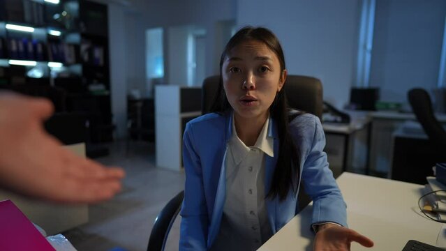 Portrait of sad stressed Asian young woman excusing herself for failures sitting in office at table with unrecognizable manager scolding gesturing. Incompetent employee apologizing for mistakes