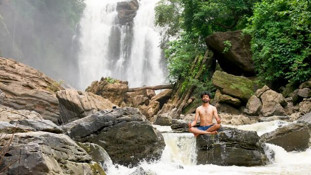 wide shot of young man in bare body on rock doing meditation with eyes closed near waterfall - concept of mindfulness, peace and lost in nature.