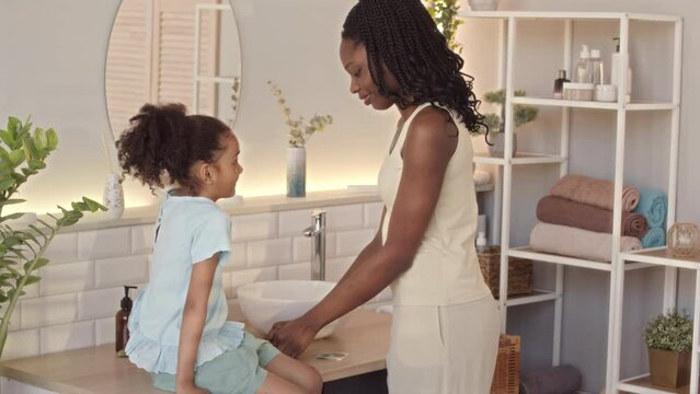 Medium slowmo of joyful young African American woman applying hydrogel eye patches on little daughter face, having fun together in bathroom doing morning routine