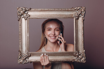 Millenial young woman blonde hair holds gilded picture frame in hands face portrait with mobile...