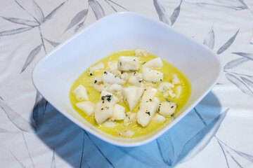 An elegant square white ceramic bowl with cuttlefish in a colorful garlic sauce on a floral patterned tablecloth. Plating and food presentation.