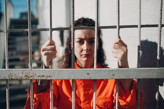Young woman in orange suit behind jail bars. Female in colorful overalls portrait. Law and justice concept