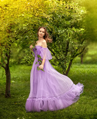 Obraz na płótnie Canvas The girl stands among the trees with lush foliage in a long lilac dress. In the background, trees, forest. Dress and hair fluttering in the wind.
