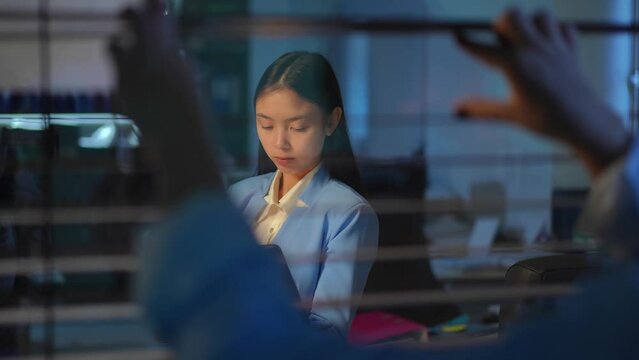 Shooting through glass of concentrated Asian woman messaging online on laptop with blurred boss peeping after employee and closing blinders. Corporate lifestyle concept