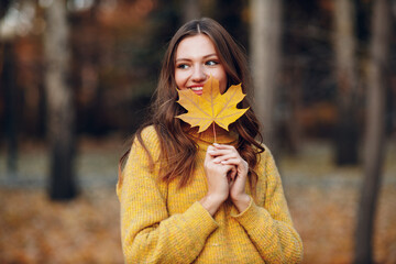 Young woman model in autumn park with yellow foliage maple leaf at face. Fall season fashion