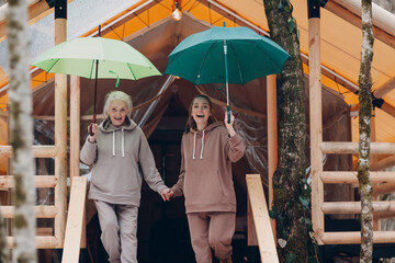 Elderly and young adult woman with umbrella at glamping camping tent. Modern vacation lifestyle...