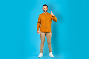 Excited european man showing thumbs up sign, standing against blue background, full body length