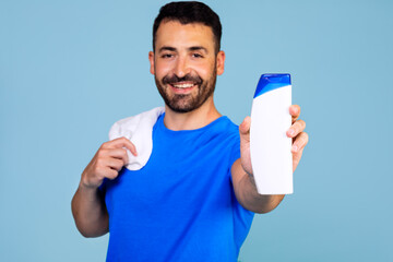 A man in a blue t-shirt and a towel holds a bottle of shampoo or shower gel on a blue background....