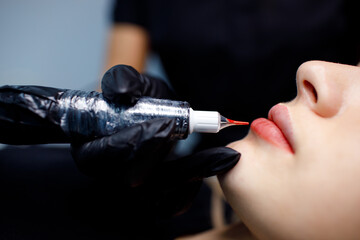 Cosmetologist applying red permanent make up tattoo on young woman lips.
