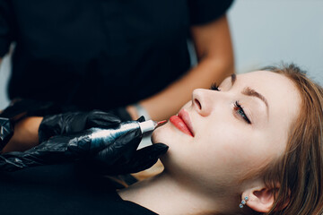 Cosmetologist applying red permanent make up tattoo on young woman lips.