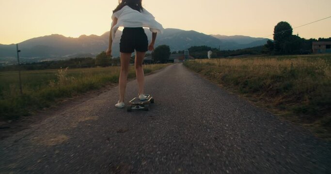 Camera following trendy girl riding on skate longboard at sunset on beautiful mountain view in countryside. Happy young stylish woman skateboarding at evening on summer vacation