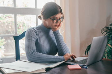 One young pretty woman studying and doing homework at home on the table. Female teenager using laptop or computer surfing the net indoor.