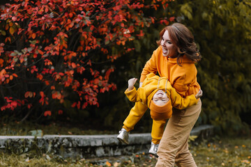 A cheerful one parent and a laughing child in warm yellow knitted sweaters are playing having fun while walking through the autumn park in nature in fall. A happy family spends time together