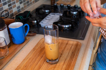 A woman hand preparing a glass of cold coffee on a wooden cutting board at kitchen