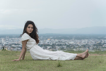 Fototapeta na wymiar woman half sitting,half lying on grass at top mountian look at camera,sky,city and nature view background. feel relax.