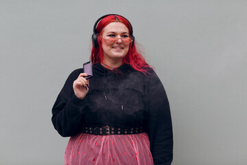 European plus size woman in headphones smoking vape electronic cigarette. Young red pink haired...