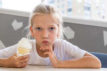 Portrait of a little hungry funny girl eating cold delicious ice cream in a waffle cup sitting in a...