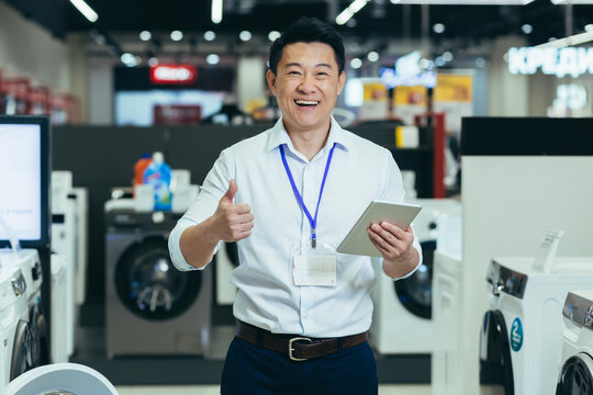 Successful sales consultant in electronics supermarket, Asian man working at home appliances store smiling and looking at camera, man holding tablet portrait and showing thumb up recommending