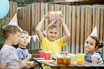 Cute funny nine year old boy celebrating his birthday with family or friends in a backyard. Birthday party. Kid wearing party hat and holding gift box.