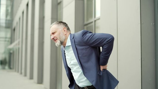 Mature business man holding his back feels back pain at work outside Spine Apin. suffering from backache spinal injury and muscle issue problem outdoor standing on a city street near office building