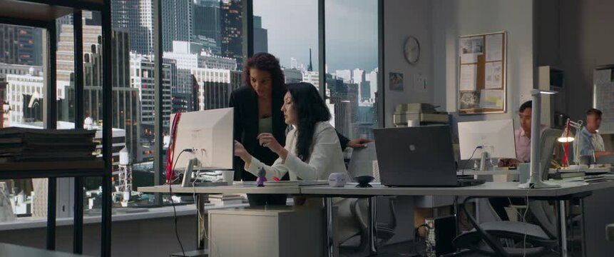 Two diverse female office employees working on something together in the modern busy office. View on a business district with skyscrapers in the background