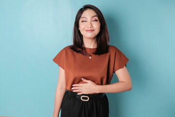 Pleased cheerful Asian woman keeps hand on belly feels full after delicious dinner stands against blue background. © Reezky