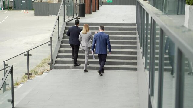 A Group Of Business People Climb Up The Stairs To An Office Building. Rear Back View A Team Of Partners Are Walking Down The Street Of Downtown. Outside. Colleagues Employees Go. Mature Man And Woman