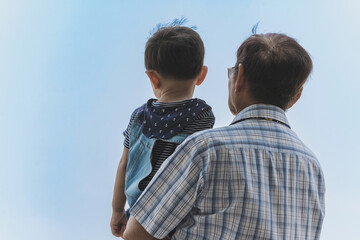 Back view of happiness Asian grandfather holding grandson with cloud and sky in background. Family stand to looking at beautiful sky together. Joy of outdoor living together. Happy family relationship