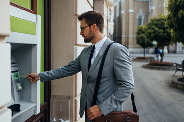 Handsome middle age businessman with eyeglasses standing on city street and using ATM machine to...