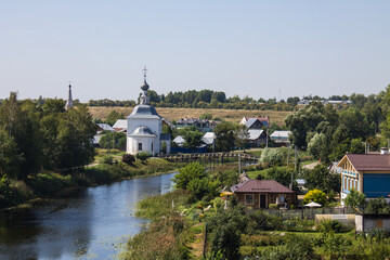Fototapeta na wymiar The white stone Church of Elijah the Prophet on the bank of the Kamenka river in Suzdal Russia on a summer day and old houses among lush green foliage