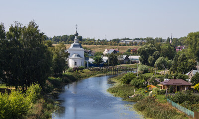 Fototapeta premium The white stone Church of Elijah the Prophet on the bank of the Kamenka river in Suzdal Russia on a summer day and old houses among lush green foliage