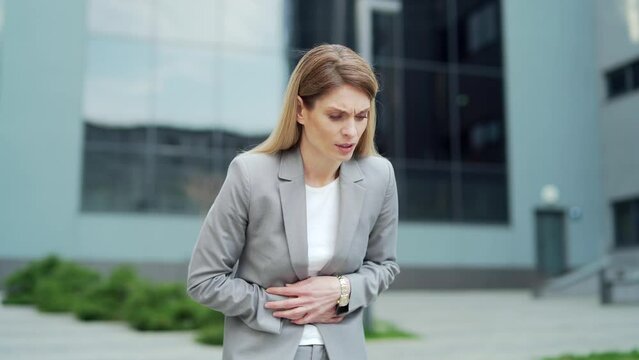 Business woman employee standing on city street with severe abdominal pain, upset stomach, diarrhea, premenstrual syndrome or poisoning. Female suffering holding stomach outside, near office building